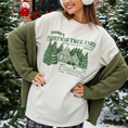 Load image into Gallery viewer, Woman wearing a Christmas-themed sweater and ear muffs in a snowy outdoor setting with trees and a building in the background.
